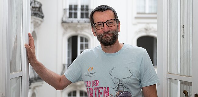 Mann mit bedrucktem T-Shirt, kurzen Haaren, Bart und Brille steht vor einer geöffneten Balkontüre. Im Hintergrund sieht man eine Häuserfront mit Fenstern