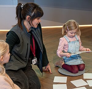 Eine junge Dame sitzt mit Kindern, die sich Bildtafeln anschauen, auf dem Boden
